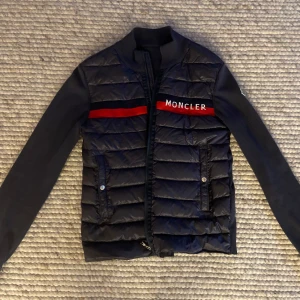 Svart Moncler jacka med röd detalj - Snygg svart jacka från Moncler med röd och svart randig detalj över bröstet och Moncler-loggan. Bra skick, skriv för övrig info!