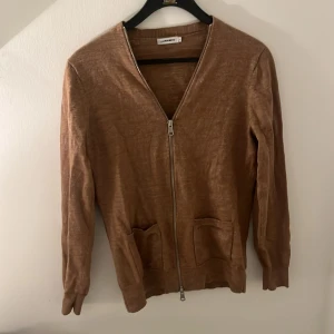 Merinoullskofta J.Lindeberg - Säljer en stilren brun kofta från J.Lindebergi merinoull med dragkedja framtill. Storlek S/XS.  Perfekt för en avslappnad och snygg look.