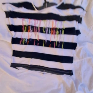 Randig topp från KappAhl - Säljer en cool randig topp från KappAhl i svart och vit med färgglada paljettdetaljer på framsidan. Toppen har korta ärmar och en avslappnad passform. Perfekt för en färgglad touch i garderoben!