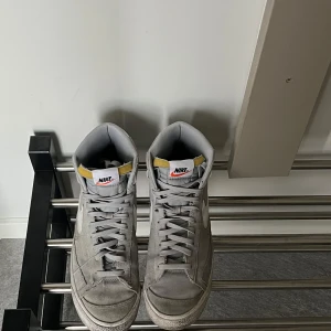 Grå Nike Blazer sneakers - Snygga grå Nike Blazer sneakers med hög modell och vit swoosh. Skorna har snörning och en klassisk design med Nike-logga på hälen. Perfekta för en stilren look.