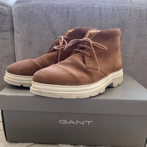 Bruna mockaboots från GANT - Snygga bruna boots i mocka från GANT med snörning och en robust vit sula. Perfekta för en stilren look. Modellen heter Brobuddy och är i färgen cognac.