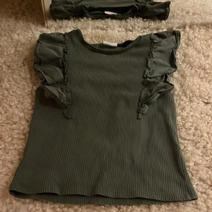 Grön ribbad topp från Kappahl - Säljer en snygg grön ribbad topp från Kappahl med volangdetaljer vid ärmarna. Perfekt för en stilren look. Toppen är ärmlös och har en bekväm passform.