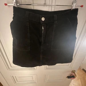 Svart manchesterkjol från Monki - Snygg svart manchesterkjol från Monki i storlek 38. Kjolen har en klassisk design med dragkedja och knapp framtill. Perfekt för att ge din outfit en trendig touch.