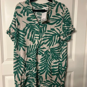 Grön och beige klänning med bladmotiv - Snygg klänning med grönt bladmotiv på beige bakgrund. Den har korta ärmar och en v-ringad hals. Perfekt för en avslappnad stil med en touch av natur.