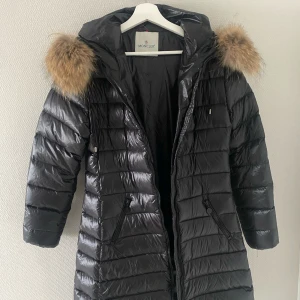 Svart dunjacka  - Snygg svart dunjacka med pälskantad huva. Jackan har en lång passform och är utrustad med dragkedja framtill samt praktiska fickor. Passar också liten large.