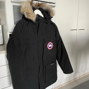 Svart parkajacka med pälsdetaljer - Säljer en svart parkajacka från Canada Goose med pälsdetaljer på huvan. Jackan har en broderad logotyp på bröstet och texten 'Expedition' på ryggen. Perfekt för kalla vinterdagar med sin långa passform och varma material.
