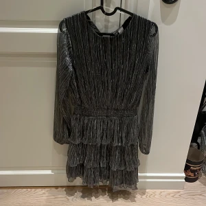 Glittrig klänning från Lindex - Säljer en glittrig klänning från Lindex i silver med långa ärmar och volangdetaljer. Perfekt för en festlig look! Klänningen har en knapp i nacken för enkel påklädning. ✨