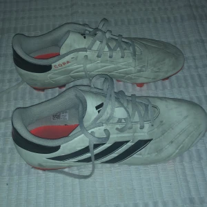 Vita Adidas Copa fotbollsskor - Säljer ett par vita Adidas Copa fotbollsskor med svarta detaljer och röda dobbar. Skorna har snörning och är perfekta för fotbollsspel på gräs. De har en klassisk design med tre svarta ränder på sidan. 