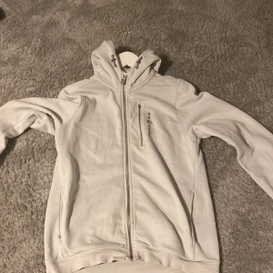 Vit hoodie från Sail Racing - Säljer en stilren vit hoodie från Sail Racing. Tröjan har en dragkedja framtill och en praktisk bröstficka med dragkedja. Perfekt för kyliga dagar med sin mjuka och bekväma passform. Huvan ger extra värme och stil. Passar både till vardags och mer avslappnade tillfällen. Bröstfickan saknar en zip därför det låga priset.