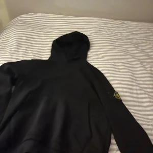 Svart hoodie från Stone Island - Säljer en stilren svart hoodie från Stone Island. Den har en klassisk design med en huva och en ikonisk logga på ärmen.