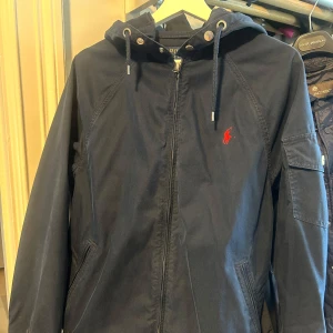 Mörkblå jacka från Polo Ralph Lauren - Snygg mörkblå jacka från Polo Ralph Lauren med huva och dragkedja. Jackan har en röd broderad logotyp på bröstet och praktiska fickor. Perfekt för en stilren look.