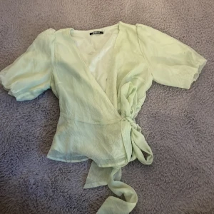 Ljusgrön omlottblus från Gina Tricot - Säljer en ljusgrön omlottblus från Gina Tricot. Blusen har korta puffärmar och ett knytband i midjan. Materialet är lätt och luftigt, perfekt för varmare dagar. Den har en v-ringad design som ger en elegant touch. Betalning sker via Swish och köparen står för frakten.🤍