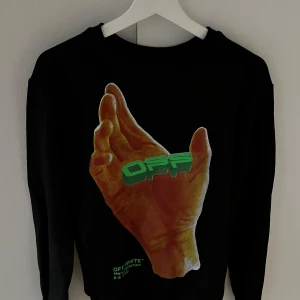 Svart tröja från Off-White - Säljer en svart tröja från Off-White med ett unikt tryck av en hand och texten 'OFF' i grönt på framsidan. Baksidan har en mindre logotyp i grönt. Tröjan har långa ärmar och en rund halsringning. Perfekt för dig som gillar streetwear!