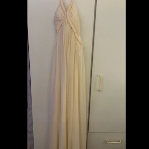 Champagnefärgad långklänning - Elegant champagnefärgad långklänning från Proms&Weddings med flätade detaljer vid halsen. Klänningen har en vacker drapering och är perfekt för speciella tillfällen. 