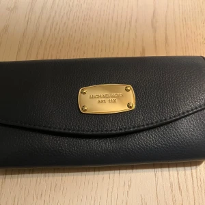 Mörk blå plånbok från Michael Kors - Mörk blå plånbok från Michael Kors i skinn med guldfärgad logotyp på framsidan. Plånboken har flera fack och en dragkedja inuti för säker förvaring. Perfekt för att hålla ordning på kort och kontanter.