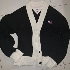 Svart och vit kofta från Tommy Hilfiger - Säljer en snygg svart och vit kofta från Tommy Hilfiger. Koftan har en klassisk design med knappar framtill och en broderad logotyp på bröstet. Den är stickad med ribbade muddar vid ärmslut och nederkant. Perfekt för kyliga dagar! Passar Storlek s-L
