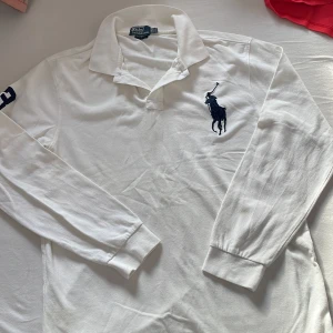 Vit långärmad pikétröja från Ralph Lauren - Säljer en klassisk vit långärmad pikétröja från Ralph Lauren med den ikoniska broderade logotypen på bröstet. Tröjan har en krage och knappar framtill, perfekt för en stilren look. Passar bra till både jeans och chinos.