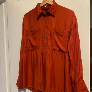 Rostfärgad skjorta från Lindex - Snygg rostfärgad skjorta från Lindex i 100% viskos.  Skjortan har långa ärmar och silklinkade känsla. Perfekt för en stilren look med knappar framtill och klassisk krage. Strl s-m