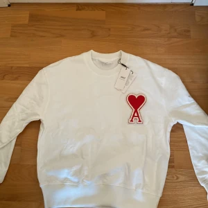 Vit sweatshirt från AMI - Säljer en stilren vit sweatshirt från AMI med ett stort rött hjärta och bokstaven A på bröstet. Tröjan har en rund halsringning och långa ärmar. Perfekt för en avslappnad och trendig look. Tröjan är i storlek S men passar också M