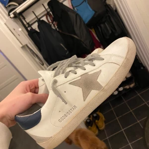 Golden goose - Snygga vita sneakers från Golden Goose med en ikonisk stjärndetalj i grått på sidan. Skorna har grå snörning och en mörkblå häl. Perfekta för en avslappnad stil. Dustbag medföljer men box är tyvärr borttappad. Skorna är i storlek 41 men är lite små i storleken. Hör av er vid fler bilder eller frågor Obs: pris kan diskuteras vid snabb affär