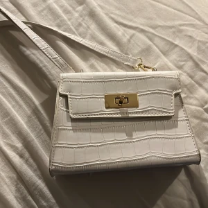 Vit axelväska - Snygg vit axelväska och gulddetaljer. Väskan har en elegant låsdetalj framtill och en justerbar axelrem. Perfekt för att ge en stilren touch till din outfit.