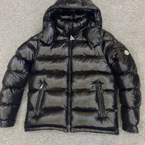 Svart dunjacka från Moncler - Snygg svart dunjacka från Moncler med huva och dragkedja. Jackan har en glansig finish och två praktiska fickor med dragkedjor. Perfekt för kyliga dagar.