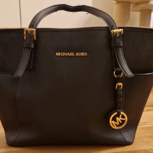 Svart handväska från Michael Kors - Snygg svart handväska från Michael Kors med gulddetaljer och logotyp. Väskan har två handtag och en dragkedja upptill. Perfekt för att bära dina dagliga nödvändigheter med stil.