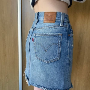 Jeanskjol från Levi's - Snygg jeanskjol från Levi's i klassisk blå denim. Kjolen har en kort längd med slitna detaljer och fransig nederkant. Perfekt för en avslappnad stil. Den har fem fickor och en knappgylf.