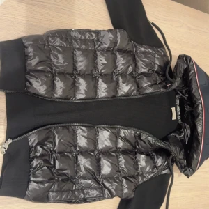 Svart cardigan från Moncler - Snygg svart cardigan från Moncler med quiltad design och dragkedja. Jackan har långa ärmar och en huva med dragsko. Perfekt för kyligare dagar med stil. Tag ingår. Storlek S/M