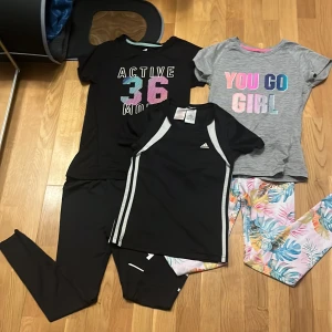 T-shirtar med tryck och träningsset - Säljer ett set med tre t-shirtar och ett träningsset. En svart t-shirt med texten 'ACTIVE 36 MODE' i färgglada bokstäver och en grå t-shirt med texten 'YOU GO GIRL'. Träningssetet från Adidas är svart med vita detaljer och består av en t-shirt och byxor.