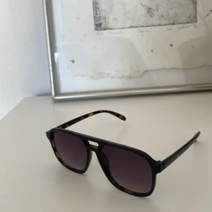 Solglasögon med leopardmönster - Snygga solglasögon med svart front och leopardmönstrade skalmar. De har en modern design med fyrkantiga linser och en stilren brygga. Perfekta för att ge din stil en extra touch.