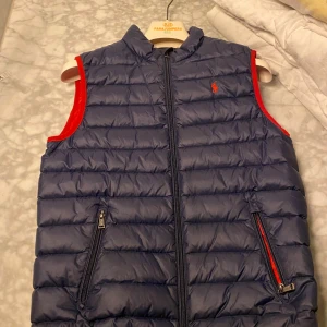 Blå dunväst från Ralph Lauren - Snygg blå dunväst från Ralph Lauren med röd insida och dragkedja. Perfekt för kyliga dagar med stil. Västen har två praktiska fickor med dragkedjor och en broderad logga på bröstet.