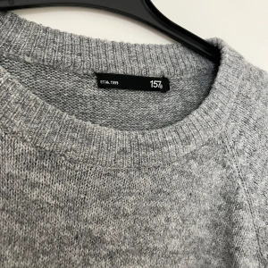 Grå stickad tröja från 157 - Mysig grå stickad tröja från 157 med rund hals och långa ärmar. Perfekt för kyliga dagar och enkel att matcha med olika outfits. Tröjan har en klassisk design med ribbade kanter vid hals, ärmslut och nederkant.