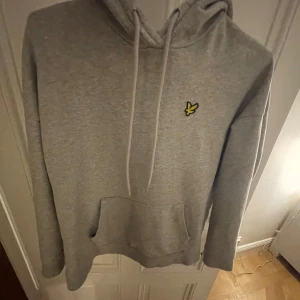 Grå hoodie från Lyle & Scott - Säljer en stilren grå hoodie från Lyle & Scott med deras ikoniska gula logga på bröstet. Tröjan har en klassisk känguruficka och justerbar huva med dragsko. Perfekt för en avslappnad look.