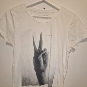 Vit t-shirt med peace tecken - Säljer en vit t-shirt med ett coolt tryck av en hand som gör ett peace-tecken och texten 'FREEDOM'. Garderobsrensning så kika gärna på övriga auktioner.