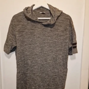 Hoodie - Säljer en grå/grön kortärmad hoodie med svarta ränder på ärmarna. Perfekt för en avslappnad stil. Den har en mjuk och bekväm känsla med en luva för extra stilpoäng.