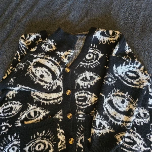 Svart kofta med ögonmönster - ALL PÄLS ROLLAS BORT INNAN FÖRSÄLJNING, kunde inte få bort allt ju för bilden, lite lat kanske! ~Säljer en unik svart kofta med ett iögonfallande ögonmönster i vitt. Koftan har knappar framtill och ribbade kanter. Perfekt för dig som vill sticka ut med en cool och annorlunda stil.