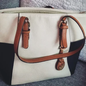 Vit och svart vintage axelväska med bruna detaljer - Elegant vintage axelväska i vitt och svart med bruna remmar och detaljer. Väskan har en praktisk dragkedja upptill och en dekorativ medaljong. Perfekt för att bära dina dagliga nödvändigheter med stil.