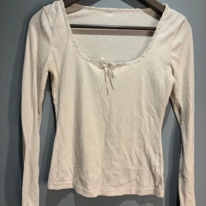 Vit ribbad långärmad topp med rosett - Säljer en söt vit ribbad topp med långa ärmar och en fin rosett vid halsringningen. Perfekt för en stilren och enkel look. Toppen har en mjuk och bekväm passform.