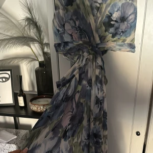 Blommig långklänning med knytband - Säljer en vacker långklänning med blommigt mönster i blå och gröna toner. Klänningen har en omlott-design på ena benet ,  midjan har öppning på båda sidorna fram och bak för extra stil. Perfekt till sommaren och midssomar ! Nypris 600kr. Endast använd på midssomar 