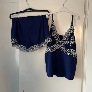 Mörkblå satinlinne med spetsdetaljer - Elegant mörkblå linne i satin med vackra vita spetsdetaljer. Linnet har smala axelband och matchande shorts med spetskant. Perfekt för en lyxig och bekväm känsla.