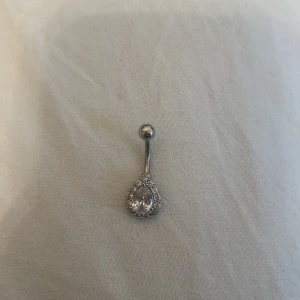 Silver navelpiercing med glittrig sten - Elegant navelpiercing i silver med en stor glittrig sten i droppform omgiven av mindre stenar. Perfekt för att ge en extra touch av glamour.