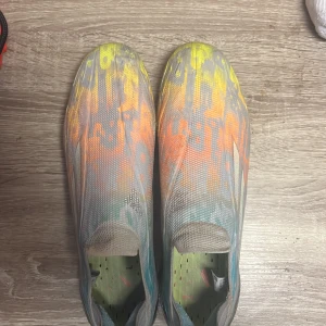 Färgglada fotbollsskor - Säljer ett par färgglada fotbollsskor med ett unikt mönster i gult, orange och blått. Skorna har en strömlinjeformad design utan snörning för en tight passform. Perfekta för att sticka ut på planen!