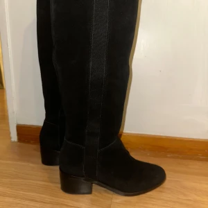 Svarta mockastövlar från Steve Madden - Snygga svarta stövlar i mocka från Steve Madden. De har en klassisk design med låg klack och är perfekta för både vardag och speciella tillfällen. Stövlarna är knähöga och har en stilren look. Stl 36. Använd endast ett fåtal gånger. Klackhöjd 3,5cm 