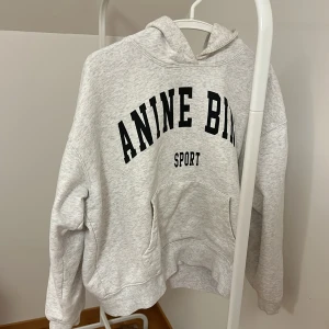 Grå hoodie från Anine Bing - Säljer en stilren grå hoodie från Anine Bing med svart text på bröstet. Tröjan har en klassisk design med huva och långa ärmar, perfekt för en avslappnad look. Den är mjuk och bekväm, idealisk för vardagsbruk.