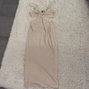 Beige axelbandslös klänning - Snygg beige axelbandslös klänning med en unik design som har en twistad detalj framtill. Perfekt för en elegant look. Klänningen är i en mjuk och bekväm polyesterblandning.