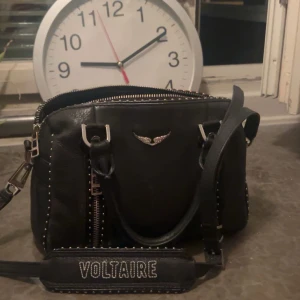 Svart handväska från Voltaire - Snygg svart handväska från Voltaire med silverdetaljer och en justerbar axelrem. Väskan har en dragkedja upptill och ett dekorativt emblem framtill. Perfekt för att bära dina dagliga nödvändigheter med stil.