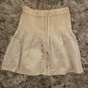 Vit kjol från Zara - Säljer en söt vit/beige kjol från Zara med smockdetaljer och knytband i midjan. Kjolen har en medellång längd och fina pärlor framtill. Perfekt för en sommaren!💗