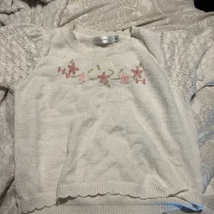 Vit stickad tröja med blommor från Knit Mix - Charmig vit stickad tröja från Knit Mix med broderade blommor i rosa och grönt på bröstet. Tröjan har korta ärmar med ett fint mönster och ribbade kanter. Perfekt för en söt och avslappnad stil. Passar st S