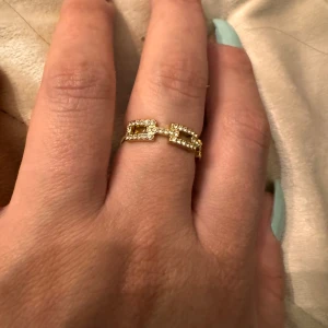 Guldfärgad ring från Pilgrim - Snygg guldfärgad ring från Pilgrim med en unik design som liknar kedjelänkar. Ringen är dekorerad med små glittrande stenar som ger den en lyxig känsla. Perfekt för att piffa upp vilken outfit som helst! 